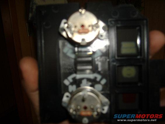 Dashboard instrument cluster with two circular gauges and three rectangular displays. A hand is holding the part