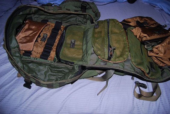 Open green and tan backpack with multiple zippered compartments and straps, laid on a blue striped surface