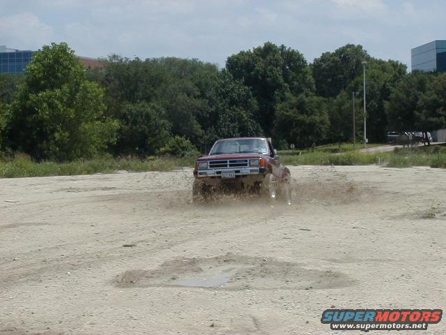 Red pickup truck kicking up mud while driving off-road. The truck appears to be from the 1980s