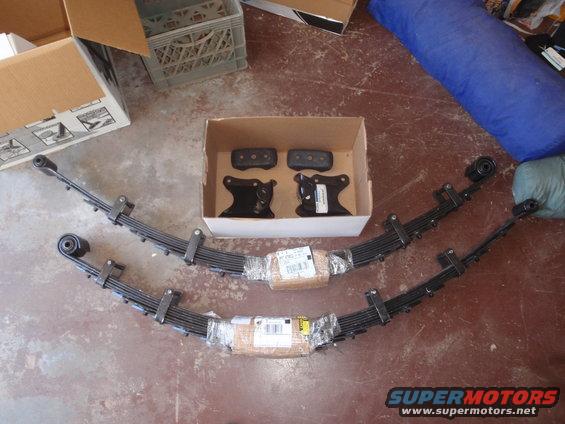 Two leaf springs and some brackets are laid out on the floor, with boxes and a blue bag in the background