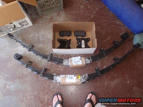 Two leaf springs and suspension brackets in a box on a concrete floor