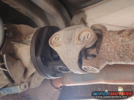 Close-up of a vehicle's driveshaft connected to a universal joint, showing rusty metal components and part of the exhaust pipe
