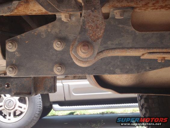 Underside of a vehicle showing rusty metal brackets and suspension components. A silver SUV is visible in the background