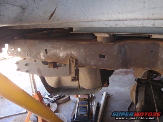 Rusty vehicle frame with suspension parts visible, supported by jack stands and a hydraulic jack