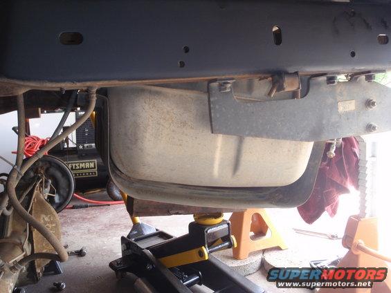 Underbody view of a vehicle's fuel tank being supported by jack stands and a hydraulic jack. An air compressor is visible in the background
