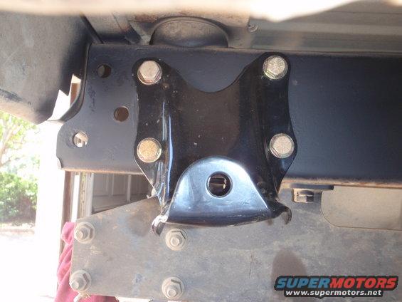 Close-up of a black metal suspension bracket with bolts, attached to a vehicle frame. Supermotors.net logo visible