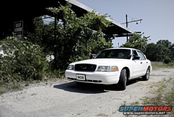 White 2002 Ford Crown Victoria parked on a gravel road near a building with overgrown bushes