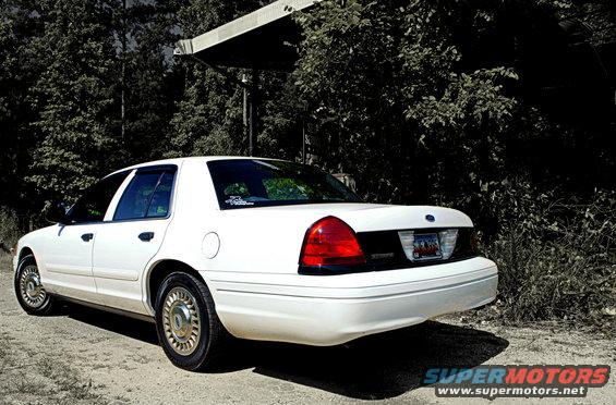 White 2002 Ford Crown Victoria parked on dirt road, trees and overhang in background