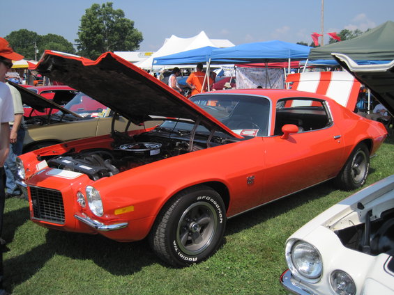 Orange 1970s muscle car with hood open, showing engine. Another car is partially visible in the foreground