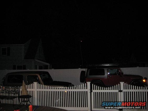 Two Jeep Wranglers parked behind a white picket fence at night. One is dark colored, the other is red and silver with a roof rack
