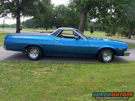 Blue 1973 Ford Ranchero parked on a paved surface next to a grassy area with trees in the background