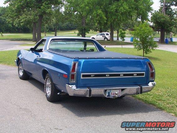 Blue Ford Ranchero parked on asphalt with a black tonneau cover. Another white SUV is visible in the background