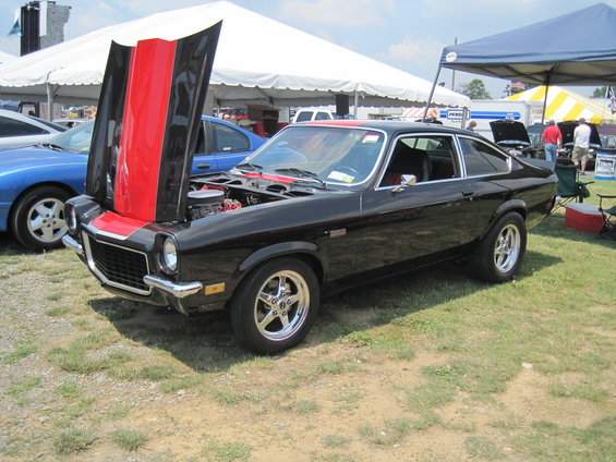 Black coupe with red and black stripes on hood, hood open showing engine. Other cars and tents in background