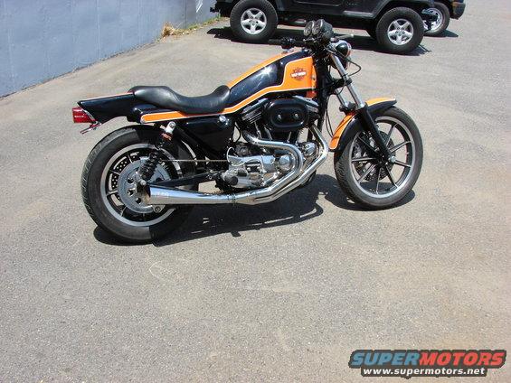 Harley-Davidson Sportster motorcycle with orange and black paint, parked near two SUVs