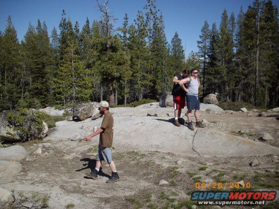 Three men stand on a rocky outcrop with pine trees in the background. A date and website are watermarked on the lower right