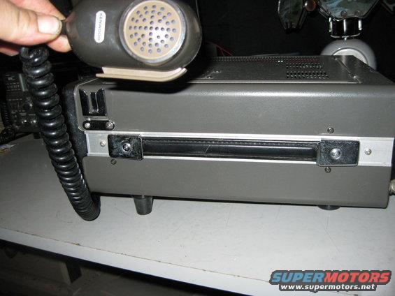 Gray radio transceiver with black coiled cord microphone held by a hand. The radio has a silver trim and a black handle