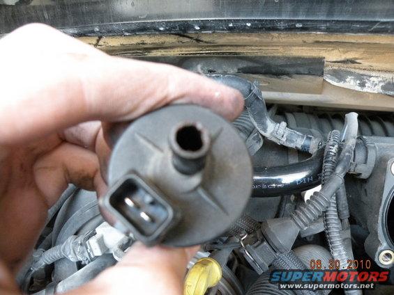 Hand holding a black automotive part with a two-pin connector and a central hole, with engine components visible in the background