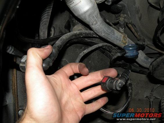 Hand holding a black fuel line connector with a red tab, attached to hoses and wiring