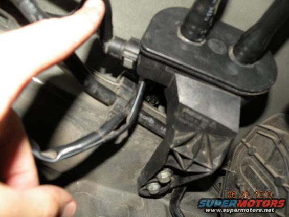 Close-up of a black plastic automotive component with attached hoses and wiring, a human finger is visible on the left