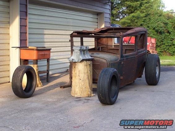 Rusty sedan body, likely a 1928 Ford Model A, on large tires, with engine parts on a log, in front of a garage