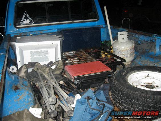 Blue pickup truck bed filled with camping gear, including a propane tank, grill, chairs, and a white cooler