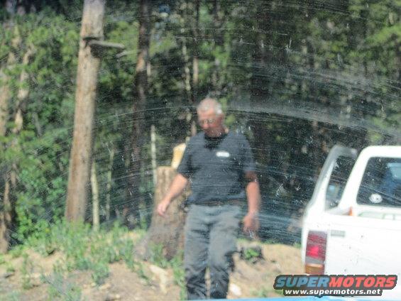 Man in black shirt and gray pants walks near a white pickup truck with open door in wooded area