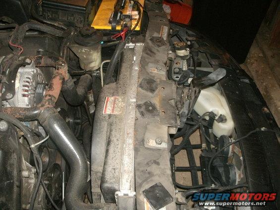 Car engine bay with radiator, alternator, hoses, and battery visible. Some labels are present