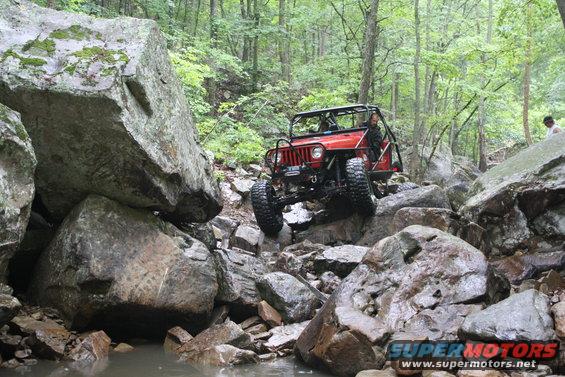 Red Jeep navigating a rocky trail with large boulders and trees