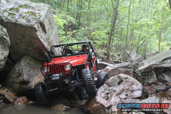 Red off-road jeep with roll cage navigating a rocky stream