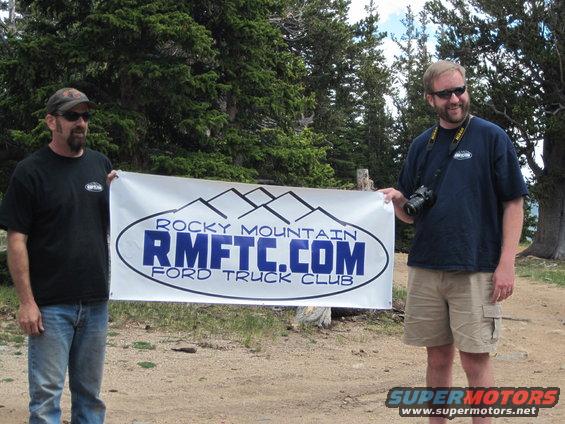Two men stand outdoors holding a banner that says "ROCKY MOUNTAIN RMFTR.COM FORD TRUCK CLUB"