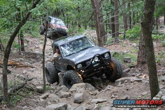 Two off-road vehicles navigate a rocky trail in a wooded area. The lead vehicle is a modified SUV with large tires
