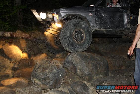 Black SUV with large tires on rocks, driver visible, headlights on