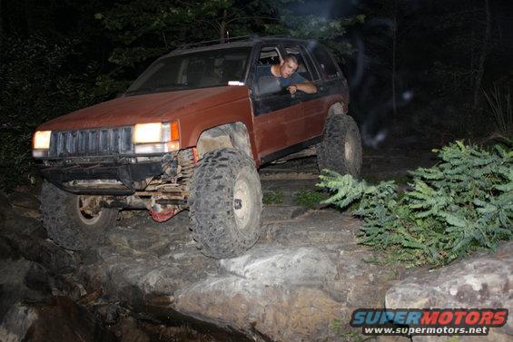 Red SUV with headlights on, driving on a dirt path at night, surrounded by trees