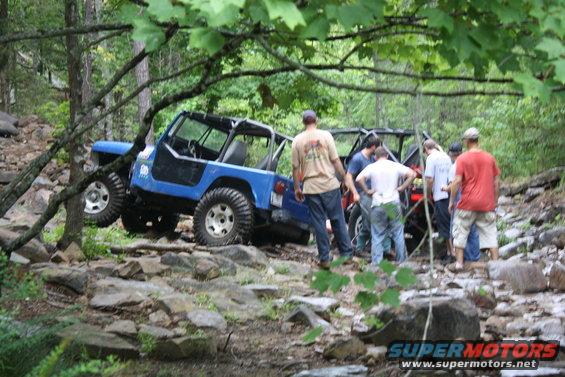 Blue Jeep Wrangler navigating rocky terrain with people gathered around