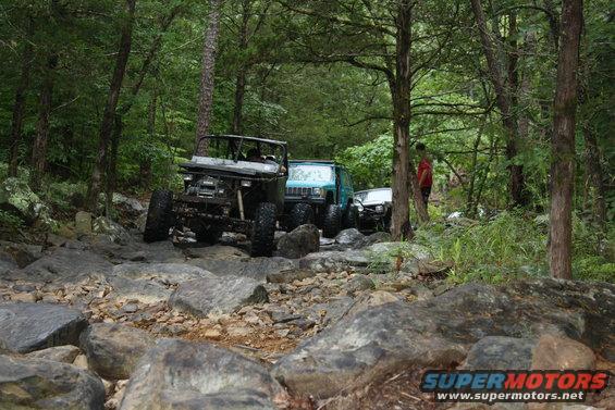 Several off-road vehicles navigate a rocky, wooded trail. One vehicle is a dark-colored Jeep-like SUV, and another is a lighter-colored SUV behind it