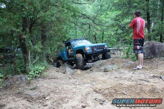 Teal Jeep Cherokee navigating a rocky, wooded trail. Another vehicle is visible in the background. A person stands watching