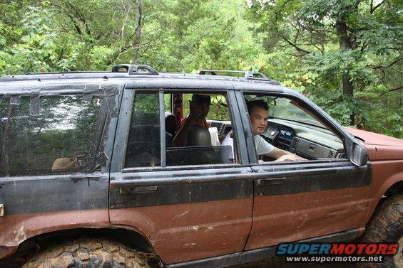 Side view of a muddy off-road SUV with two people inside, one driving. The vehicle has a two-tone paint job and roof rack