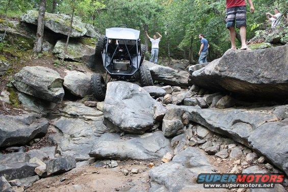 Off-road buggy with roll cage climbing over large rocks on a trail. People watch from the rocks