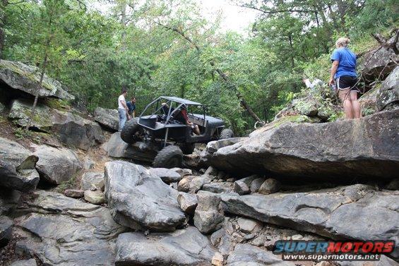 Black off-road buggy with a roll cage navigating a rocky trail, with people watching