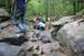 1984 Ford Bronco II - Multiple off-road vehicles tackle a challenging rocky path in a wooded area, with a person observing from large boulders.