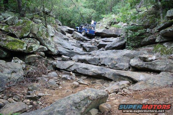 Blue off-road vehicle on a steep, rocky trail with trees and mossy rocks