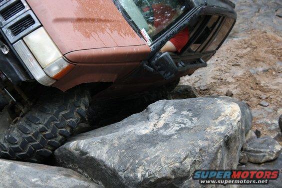 Brown SUV tilted on large rocks, driver wearing red shirt visible through window