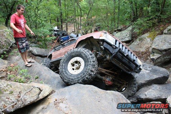 Brown 1984 Ford Bronco II tilted on large rocks, front grille visible, with a man standing nearby