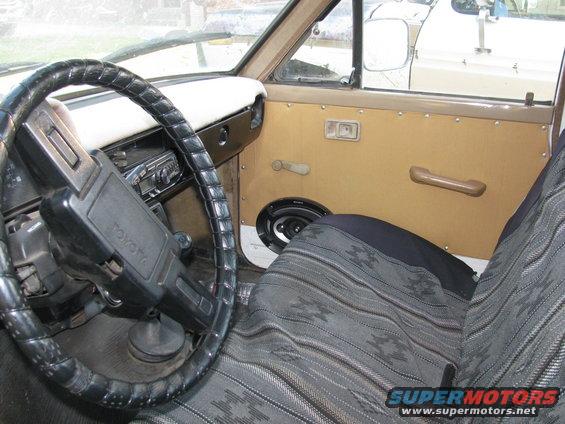 Interior of an old Toyota truck, showing the steering wheel with 'Toyota' logo, patterned seat covers, and a speaker on the door panel. Another vehicle is visible through the window