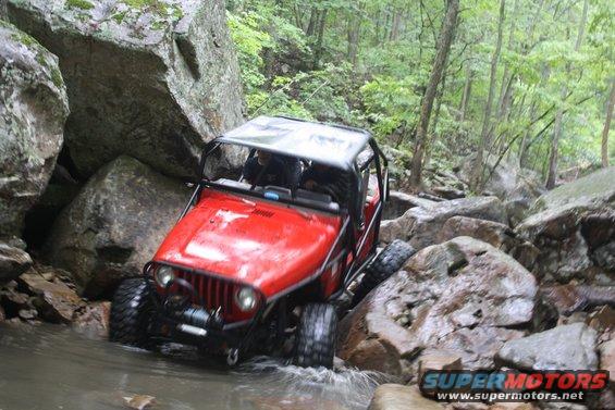 Red Jeep Wrangler crawling over rocks in a stream, trees and rocks all around
