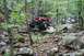 1984 Ford Bronco II - A red off-road vehicle navigates a challenging rocky trail through a wooded area.