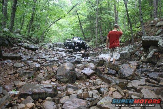 Off-road vehicle driving through a rocky, wooded area. Person in red shirt watches