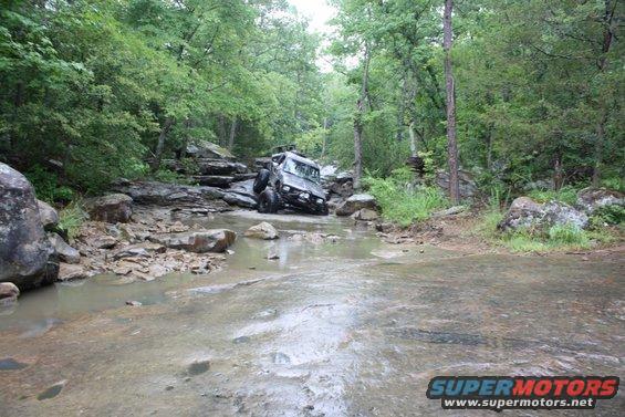 1984 Ford Bronco II driving through shallow rocky water in a wooded area