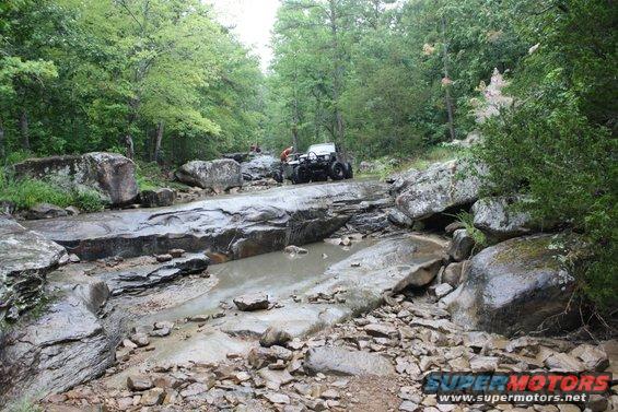 White off-road vehicle navigates rocky stream bed with trees and boulders