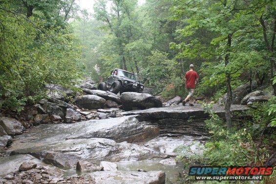 Jeep-like vehicle crawling over large rocks in a stream bed with a person walking nearby